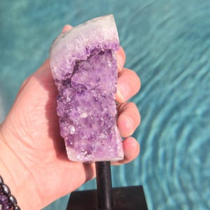 May include: A vibrant amethyst crystal cluster, showcasing a gradient of purple hues, is mounted on a black metal stand. The crystal's surface is textured, with a white edge. The background is a blurred turquoise water.