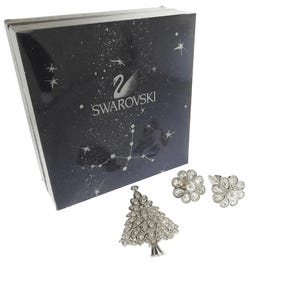 May include: A jewelry set featuring a silver-toned Christmas tree brooch and a pair of flower-shaped earrings with clear crystals and faux pearls. The set is displayed next to a black box with the Swarovski logo and a starry design.
