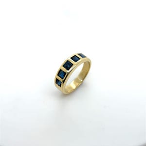 May include: A gold-colored ring featuring five square, deep blue gemstones set in a row. The ring has a smooth, polished band and a classic design. The gemstones are evenly spaced and add a pop of color to the ring.