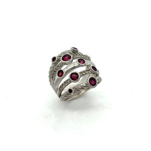 May include: A multi-strand silver ring featuring a textured, twisted rope design. The band is accented with several round, deep pinkish-red gemstones set in smooth bezels, interspersed with small, clear, sparkling stones along the metal strands.