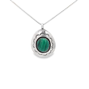 May include: A silver pendant necklace featuring a large, oval-shaped pendant with a scalloped edge. The pendant holds a round, vibrant green malachite stone. The necklace chain is delicate and silver.
