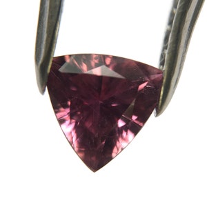 Puede incluir: Una piedra preciosa triangular facetada de un tono rosa intenso. La piedra preciosa está sujeta por unas pinzas.