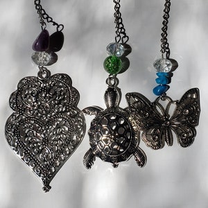 May include: Three silver tea infusers with different charms. The first infuser has a heart-shaped charm, the second has a turtle charm, and the third has a butterfly charm. The charms are attached to chains with beads.