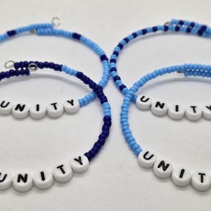 May include: Three blue and white beaded bracelets with the word "UNITY" spelled out in white beads with black lettering. The bracelets are layered on top of each other.