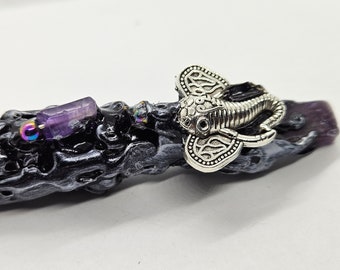 May include: A black and silver wand with a purple crystal and a silver elephant charm. The wand is decorated with a swirling pattern.