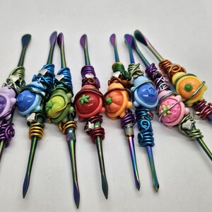 May include: A collection of colorful dab tools with decorative ends. Each tool features a different fruit or animal design, including grapes, turtles, and strawberries. The metal tools have a rainbow finish, and the decorative ends are wrapped with wire.