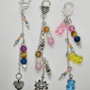 May include: Three keychains with silver-tone clasps and charms. Each keychain features a different design: one with heart and bead accents, one with a sun charm and beads, and one with gummy bear and bead accents.