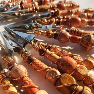 May include: A close-up of a group of hair clips with copper wire wrapped around wooden beads. The beads are various shades of brown, orange, and red, and some have intricate patterns.
