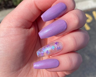 Op de afbeelding: Close-up van een hand met lange, lichtpaarse nagels met een glinsterende afwerking. Sommige nagels hebben een transparante laag met iriserende vlinderaccenten.