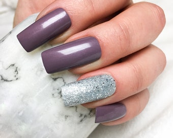 Op de afbeelding: Close-up van een hand met vier nagels die in een mauve tint zijn gelakt. De ringvingernagel heeft een zilveren glitterlak.