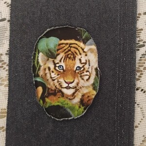 May include: A rectangular piece of dark charcoal denim fabric featuring a central oval-shaped cutout. Within this cutout is a printed image of a tiger cub with orange and black stripes, blue eyes, and green foliage surrounding its face.