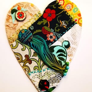 May include: A heart-shaped fabric collage featuring layered textiles including beige lace, black floral print, yellow patterned fabric, and green scrollwork. Embellishments include a teal tassel, decorative buttons, green beads, and embroidery.