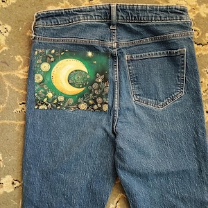 May include: Blue denim jeans featuring a rectangular patch on the left back pocket. The patch displays a yellow crescent moon surrounded by green foliage, swirling patterns, and small gold stars against a dark green and black background.