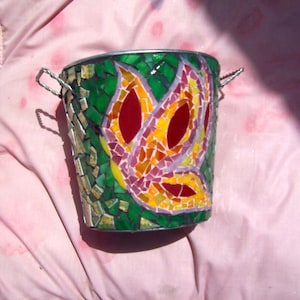 May include: A mosaic bucket with a floral design. The bucket is made of small, colorful glass tiles in shades of green, red, orange, yellow, and purple. It has a silver metal rim and wire handles. The floral design is centered on the front.