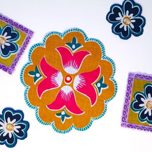 May include: A collection of five fabric pieces including two square coasters with purple lace trim, two small dark blue flower-shaped cutouts, and one large circular mustard yellow mat featuring a central pink and teal floral pattern.