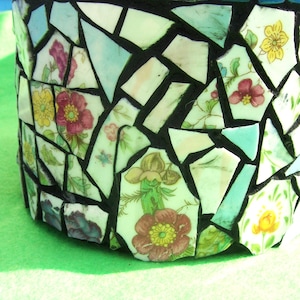 May include: A mosaic jar with a wooden lid and blue tile trim. The jar's body is made of broken ceramic pieces in various shapes, featuring floral patterns in red, yellow, and green against a white background.
