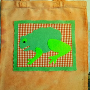 May include: A peach-coloured canvas tote bag featuring a rectangular patch with a white and orange gingham background. A green felt frog appliqué with a bright green leg and a black and white eye is centred on the patch, framed by a green border.