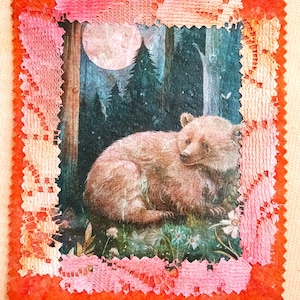 May include: A rectangular fabric patch featuring a central illustration of a brown bear resting in a forest at night under a full moon. The image is bordered by a pink and orange lace trim with a zigzag-cut edge on a textured orange background.