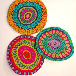 May include: Three circular fabric coasters featuring intricate, colorful mandala patterns. The top coaster displays orange, teal, and pink hues. The bottom left coaster features bright pink, lime green, and red. The right coaster shows pink, teal, and yellow.
