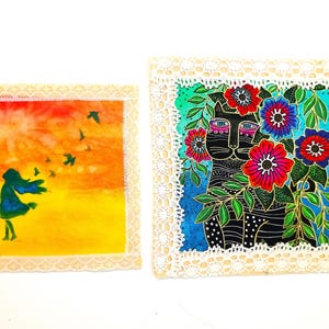May include: Two square fabric patches with white lace trim. The left patch features a silhouette of a person against a yellow and orange sunset with birds. The right patch displays a black cat surrounded by vibrant red, blue, and purple flowers.