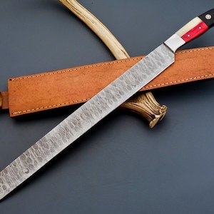 May include: A long, silver, Damascus steel knife with a red, white, and black handle. The knife is in a brown leather sheath and is resting on a piece of antler.