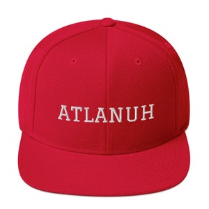 May include: A red baseball cap with a flat brim. The cap features the embroidered text "ATLANUH" in white block letters across the front. The hat has a structured fit and a rounded crown.