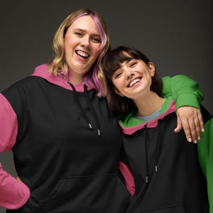 May include: Two people wearing black hoodies with color block sleeves and hood linings. One hoodie has pink accents, the other has green. Both have drawstrings and a front pocket.