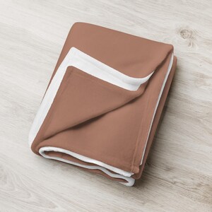 May include: A soft, plush throw blanket in a warm terracotta color with a white border. The blanket is folded in half and is lying on a light wood surface.