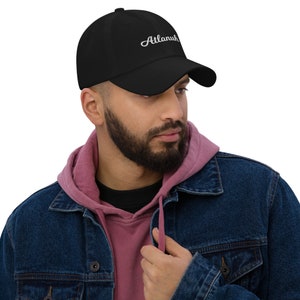 May include: A black baseball cap with a white embroidered cursive script that reads "Atlanuh".