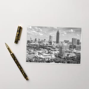 May include: Black and white postcard featuring an aerial view of the Atlanta skyline with the text "ATLANTA" printed in white.