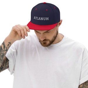 May include: A navy blue and red baseball cap with the word "ATLANUH" embroidered in white. The cap has a flat brim and a structured fit. The person in the image is wearing a white t-shirt.