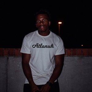 May include: A person wearing a white t-shirt with the word "Atlanuh" printed in black cursive lettering. The person is standing in front of a brick wall at night.