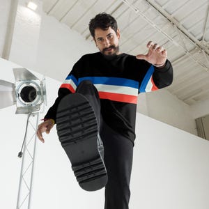 May include: A man wearing a black, blue, white, and red striped sweater with black pants and black shoes. The man is standing on a white surface and is looking at the camera.