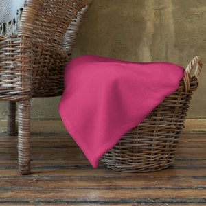 May include: A pink fabric throw draped over a wicker basket with a woven handle. The basket is sitting on a wooden floor.