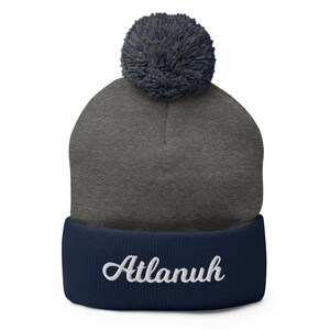 May include: A gray and navy blue beanie with a pom pom on top. The beanie has the word "Atlanuh" embroidered on the cuff in white.