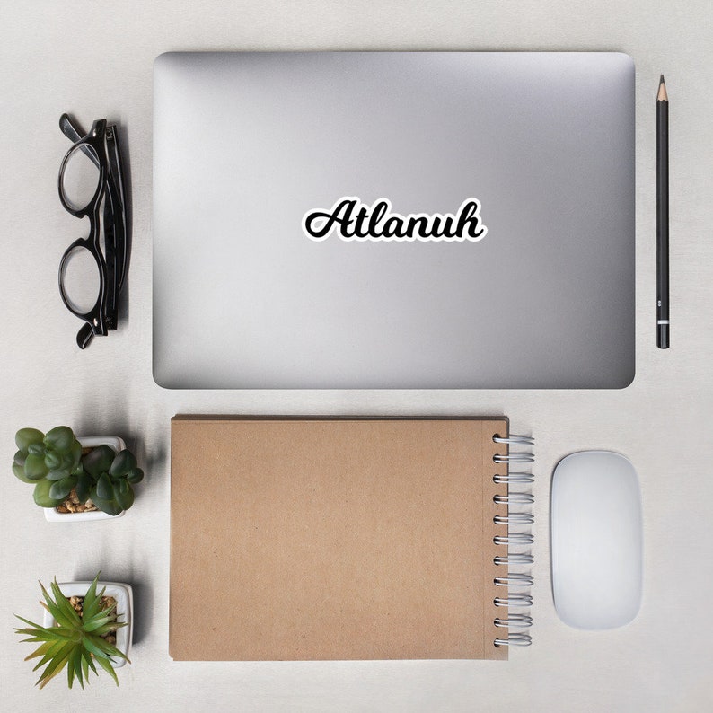 May include: A silver laptop computer with a white sticker that says "Attanuh" on the lid. The laptop is on a white surface with a brown notebook, a white mouse, and a pencil.