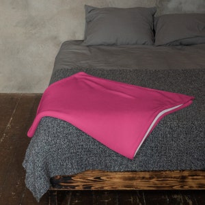 May include: A pink throw blanket on a gray bed with a wooden frame. The blanket is folded in half and has a white border.