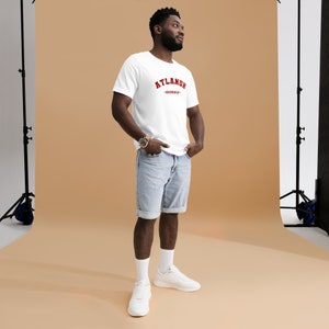 May include: A white t-shirt with red text that reads "ATLANTA - GEORGIA". The person wearing the shirt is standing in a studio setting with a light brown background.