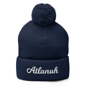 May include: Navy blue knit beanie with a white pom pom on top and the word "Atlanuh" embroidered in white on the cuff.
