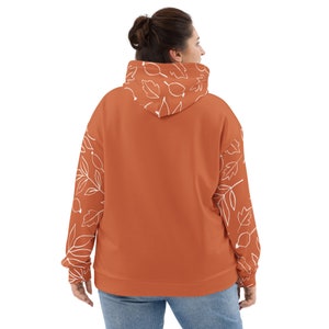 May include: A rust-colored hoodie with white leaf and berry designs on the sleeves and hood. The hoodie has a relaxed fit and a drawstring hood.