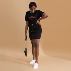 May include: A woman wearing a black t-shirt with the text "ATLANUH - GEORGIA" in red lettering. She is also wearing a black skirt and white sneakers. She is holding a black purse with a gold chain.