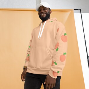May include: A peach-colored hoodie with a white drawstring and a pattern of peaches on the sleeves. The model is wearing a white baseball cap.