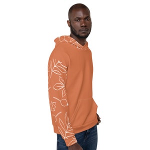 May include: A terracotta orange hoodie with a white floral pattern on the left sleeve. The hoodie has a drawstring hood and a kangaroo pocket.