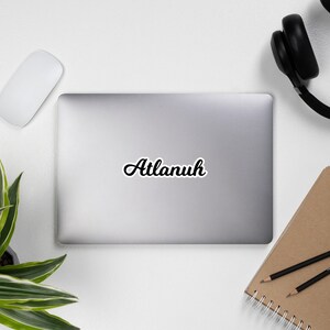 May include: A black and white sticker with the word "Atlanuh" written in a cursive font on a silver laptop.