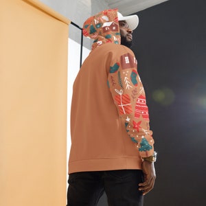 May include: A brown hoodie with a Christmas-themed print on the sleeves. The print features houses, trees, gifts, and other holiday elements. The hoodie has a hood and a white drawstring.