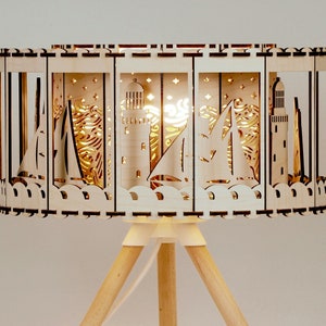 May include: A wooden nautical-themed lamp with a tripod base. The lamp shade features cut-out designs of sailboats, lighthouses, and waves. The light shines through the cutouts, creating a warm glow. The lamp has a white base and wooden legs.