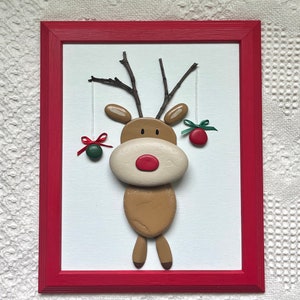 May include: A framed Christmas decoration depicting a reindeer crafted from painted stones. The reindeer has a red nose, twig antlers, and hanging ornaments. The red frame surrounds a white background, creating a festive display.