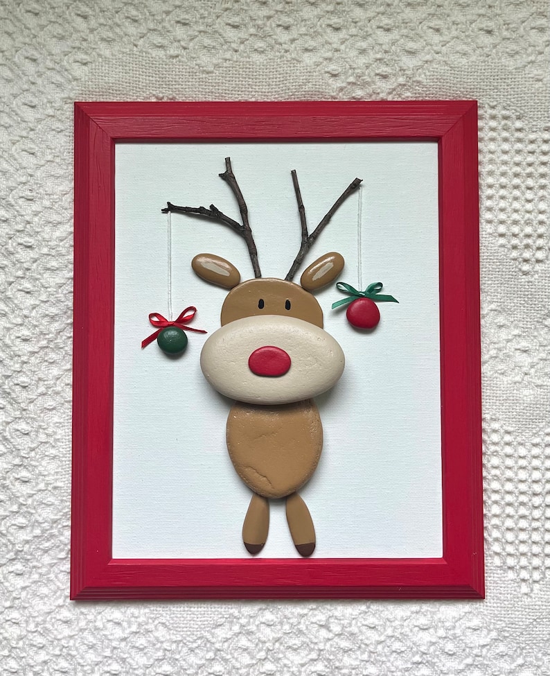 May include: A framed Christmas decoration depicting a reindeer crafted from painted stones. The reindeer has a red nose, twig antlers, and hanging ornaments. The red frame surrounds a white background, creating a festive display.