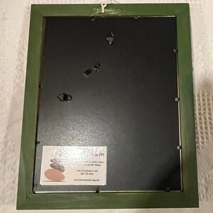 May include: The reverse of a framed artwork with a green painted wooden frame. The artwork has a black backing and a small white label with text and a logo. The frame has a wire for hanging.
