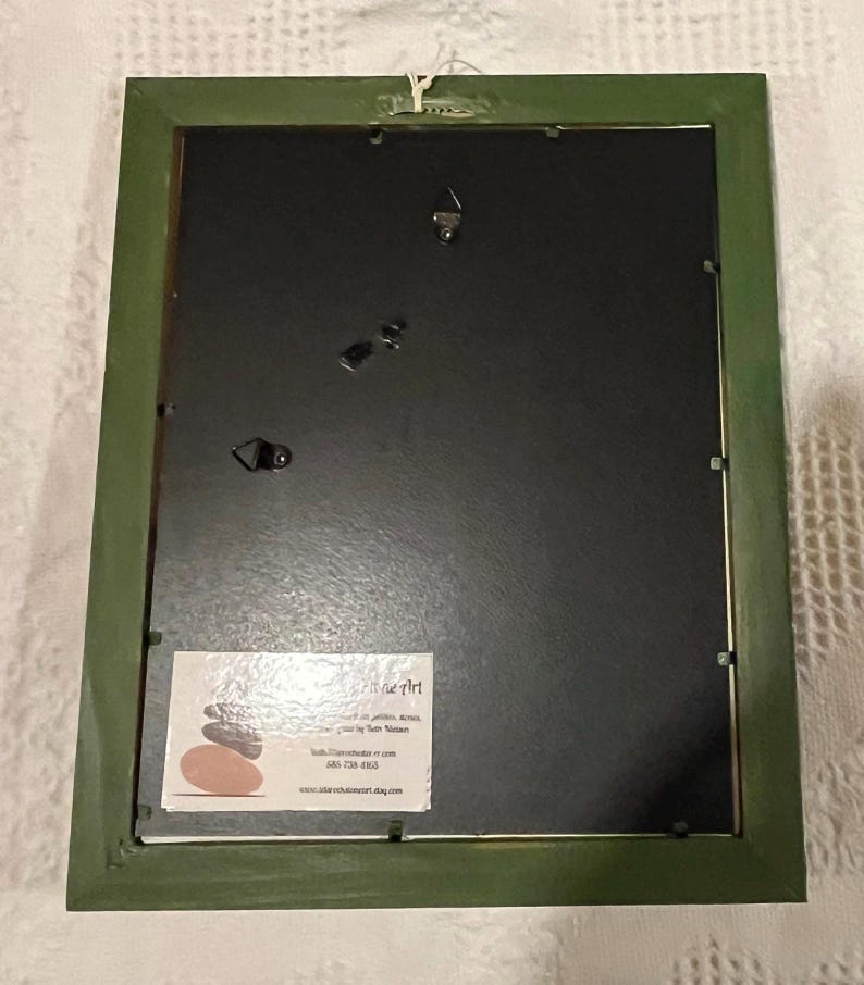May include: The reverse of a framed artwork with a green painted wooden frame. The artwork has a black backing and a small white label with text and a logo. The frame has a wire for hanging.
